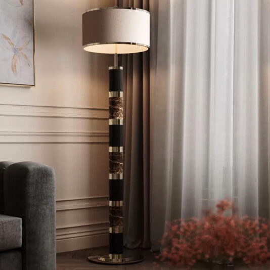 The Luxury Marble Floor Lamp, featuring a beige lampshade and a black and gold marble-patterned base, stands next to a gray sofa. Positioned near a window with sheer white curtains and complemented by a pink floral arrangement, this lamp enhances the cozy and elegant ambiance of the living room.
