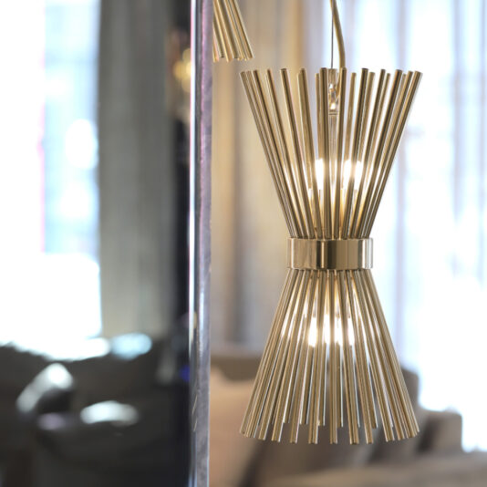 Modern Gold Plated Pendant Light A Modern Gold Plated Pendant Light with a brass finish and thin, vertical rods radiating from a central band is suspended from the ceiling. The light is illuminated, and the background reveals a blurred indoor setting with a window and furniture.