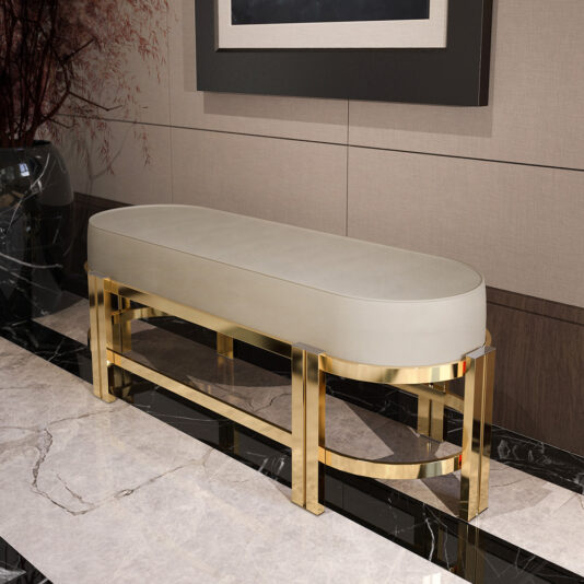 Modern Oval Leather Bench A Modern Oval Leather Bench featuring a cream cushion is positioned against a beige and wooden wall. This elegant bench boasts a shiny, gold-colored metallic frame and stands on a polished marble floor adorned with black and white patterns. A dark painted wall is visible behind the bench, adding to the contemporary ambiance.