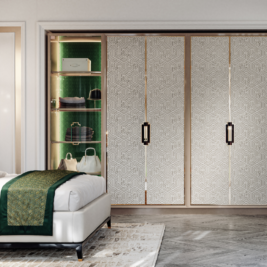 A stylish bedroom features a white bed with a green and gold-patterned throw. Behind it is a wardrobe with sliding doors, showcasing an illuminated section displaying designer bags and hats—truly a statement piece. The wood flooring complements the patterned rug partially visible beneath.