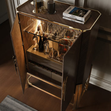 Veneer Drinks Cabinet With Feather Handle The Veneer Drinks Cabinet With Feather Handle, adorned with luxurious wooden veneer and gold accents, displays an assortment of glassware, bottles, and an ice bucket when its doors are open. Books and a candle rest on top of the cabinet, which features a distinctive feather handle. The piece stands on a gold frame positioned on a brown floor adjacent to a wall.