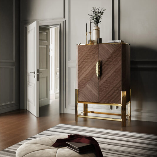 A modern, elegant interior boasts the Veneer Drinks Cabinet With Feather Handle, accented in gold. Complementing it are a plant and dark-toned candles on top, all set against a grey wall beside an open white door. Completing the sophisticated look are a striped rug and a beige cushioned seat with a maroon throw.