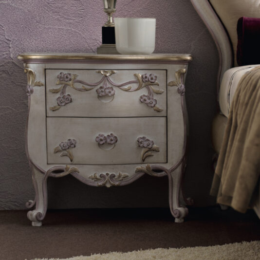 The Carved Floral Italian Bedside Cabinet is a vintage-style nightstand with ornate detailing and two drawers. It features carved floral accents and is painted in a light pastel color with gold highlights. The cabinet stands beside a bed with a beige headboard and bedding.