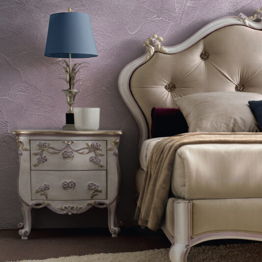 A luxurious bedroom setup featuring the Carved Floral Italian Bedside Cabinet adorned with intricate vintage-style details. On the cabinet sits a blue lampshade atop an elegant silver lamp base, accompanied by a white mug. The upholstered headboard and bed, draped in plush pillows and blankets, enhance the opulent feel.