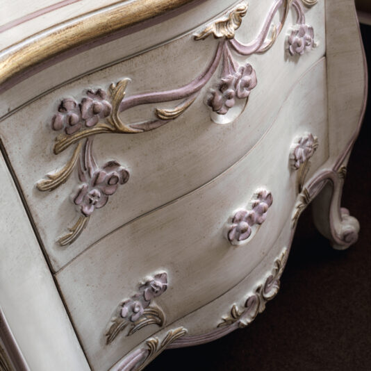 Close-up of the Carved Floral Italian Bedside Cabinet featuring gold and pink floral carvings on its drawers. This ornate vintage dresser has a distressed white finish, curved legs, and a decorative design, evoking a shabby chic or antique style.