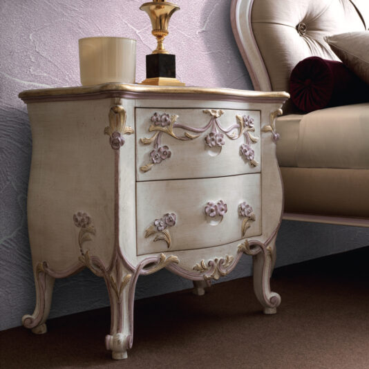 A vintage Carved Floral Italian Bedside Cabinet stands beside a beige upholstered bed. This nightstand features two drawers decorated with intricate floral designs. A candle and a gold lamp sit atop the cabinet, set against textured light purple walls in the background.