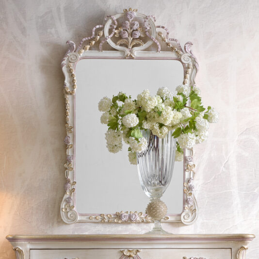 A Carved Floral Italian Wall Mirror with an ornate, vintage-style intricate frame hangs on a textured wall. In front of the mirror, there's a glass vase filled with white, round flowers, creating an elegant and harmonious display.