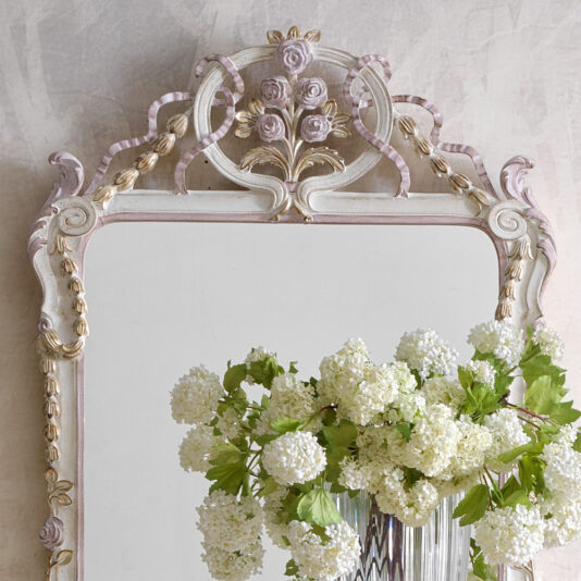 Carved Floral Italian Wall Mirror The Carved Floral Italian Wall Mirror, with its vintage-style frame adorned with ornate rose carvings at the top, provides a stunning focal point. In front of the intricately designed mirror stands a clear vase holding blooming white hydrangea flowers, set against a light, textured wall.