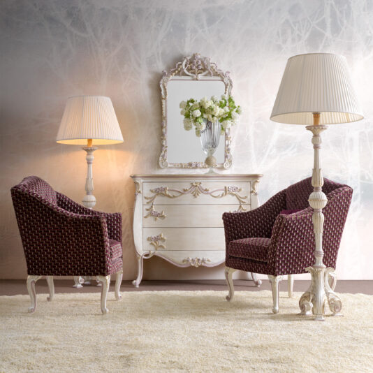Carved Floral Italian Wall Mirror A cozy living room setup featuring two vintage armchairs with maroon patterned fabric, a classic cream-colored dresser with ornate detailing, and a large Carved Floral Italian Wall Mirror above it. Beside each chair stands a tall floor lamp with a pleated shade, casting a warm glow.