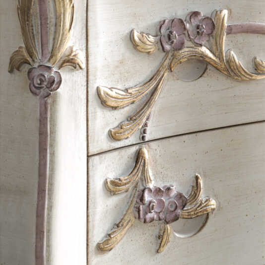 Carved Italian Chest Of Drawers Close-up of the Carved Italian Chest Of Drawers. The detail showcases ornate floral and leaf designs in soft pastel shades of pink, gold, and cream. The surface features an antique, distressed finish, giving it a vintage, elegant appearance.
