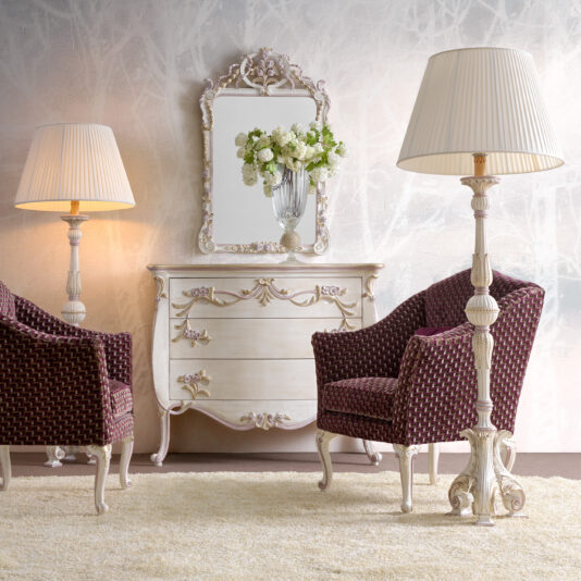 A stylish room showcases a vintage white dresser with intricate floral details and a matching ornate mirror. Two burgundy armchairs upholstered in patterned fabric flank the dresser. Tall, elegant Carved Italian Floor Lamps stand beside the chairs on a beige carpet, casting a warm glow through pleated shades.