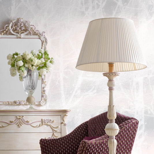Carved Italian Floor Lamp An elegant room features a vintage white dresser with ornate detailing and a large framed mirror. A glass vase filled with white flowers sits atop the dresser. Beside a maroon-patterned armchair stands a cream-colored table lamp with a pleated shade next to the Carved Italian Floor Lamp, adding an exquisite touch to the space.