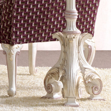A vintage-style table with an intricately carved white pedestal base on a textured beige rug. Surrounding the table are maroon cushioned chairs with a checkered pattern, all illuminated by the **Carved Italian Floor Lamp**. The floor is a warm brown color, and the background features a light pink wall.