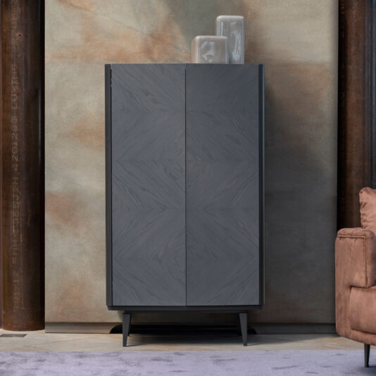 The Contemporary Ash Veneer Tall Cabinet, featuring a sleek dark grey finish and wood grain pattern, stands elegantly on four tapered legs. Two glass vases adorn its top. Positioned against a textured concrete wall near a brown cushioned chair, the cabinet rests on a light purple carpet below.