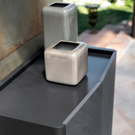 Contemporary Ash Veneer Tall Cabinet Two square, grey-tinted ceramic vases of different sizes sit on a sleek, dark grey modern table. Behind them, the Contemporary Ash Veneer Tall Cabinet subtly peeks into view amidst the softly blurred background showing outdoor greenery and a stone pathway. The overall aesthetic is minimalist and contemporary.