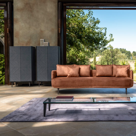 Contemporary Ash Veneer Tall Cabinet A modern living room scene featuring a brown sofa with multiple cushions, a glass coffee table with a book on it, and two Contemporary Ash Veneer Tall Cabinets in the background. Large open windows reveal lush green trees and a blue sky outside.