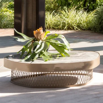 Contemporary Low Marble Coffee Table A Contemporary Low Marble Coffee Table with a mesh base and glossy surface sits outdoors, adorned with a large leafy plant on top, surrounded by a lush garden.