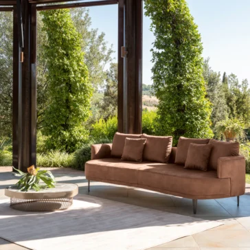 Contemporary Low Marble Coffee Table A modern outdoor seating area includes a plush-cushioned brown sofa under a wooden pergola. The Contemporary Low Marble Coffee Table with a plant rests on a light rug, surrounded by lush greenery and distant hills.