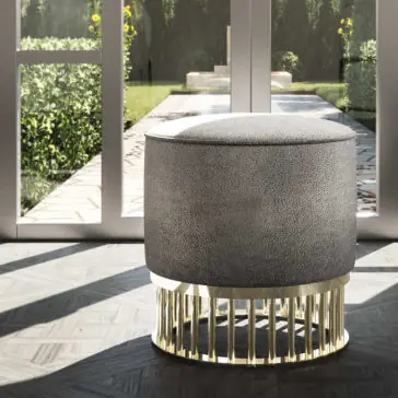 Contemporary Pouffe With Brass Base A Contemporary Pouffe With Brass Base, modern and cylindrical in form, sits in a sunlit room. Behind it, large glass doors open to a green garden pathway. The room boasts a light, textured floor and embodies a minimalist design.