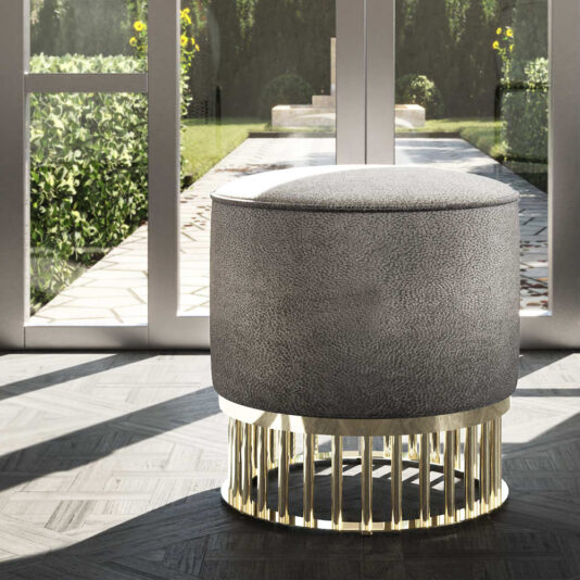 A Contemporary Pouffe With Brass Base, modern and cylindrical in form, sits in a sunlit room. Behind it, large glass doors open to a green garden pathway. The room boasts a light, textured floor and embodies a minimalist design.