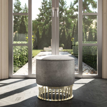 Contemporary Pouffe With Brass Base The **Contemporary Pouffe With Brass Base**, featuring a textured gray surface, is positioned before large glass doors. These doors open to a sunlit garden adorned with greenery and trees, letting sunlight flood the room and create soft shadows on the floor.