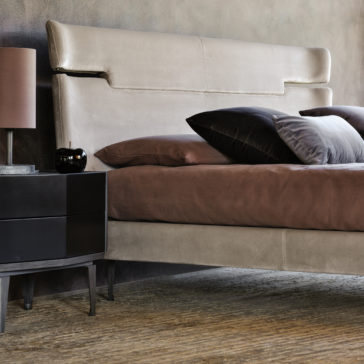 A contemporary bedroom features a taupe Contemporary Upholstered Velvet Bed adorned with brown bedding and various pillows. A black nightstand with cone-shaped legs holds a matching black table lamp and small decor items. The room showcases neutral tones and minimalist style, complete with a textured rug under the bed.