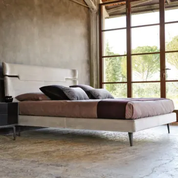 A contemporary bedroom features the Contemporary Upholstered Velvet Bed with a light beige upholstered velvet headboard and frame. The bed is dressed in brown and purple bedding complemented by several pillows. A large window with a serene outdoor view illuminates the room with natural light.