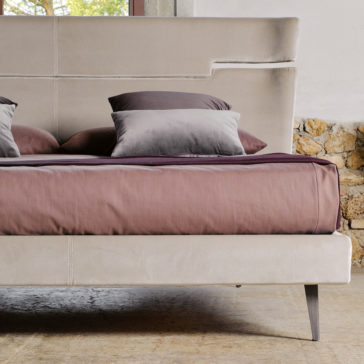 A modern bedroom setup featuring a Contemporary Upholstered Velvet Bed in light beige, accessorized with two grey gradient pillows and a purple blanket. The headboard showcases a distinctive cut-out design. In the background, a brick wall and window offer a view of outdoor greenery.