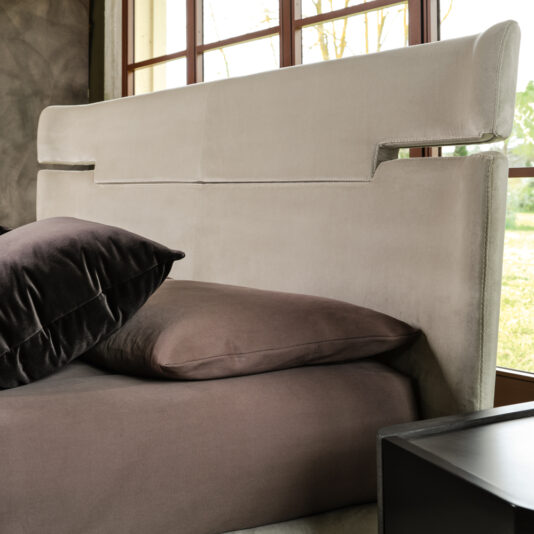 A modern bedroom showcases a luxurious Contemporary Upholstered Velvet Bed, complete with a sleek beige headboard. The bed is adorned with brown bedding and two large pillows—one brown and one black—propped against the headboard. A window offering a scenic outdoor view serves as the backdrop.