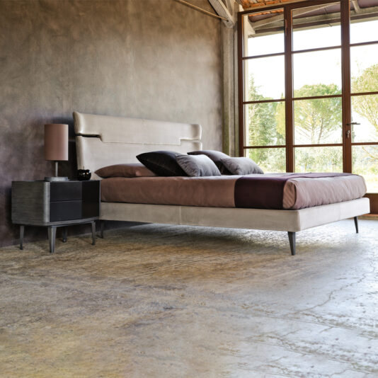 A modern bedroom with a minimalistic design. The space features the Contemporary Upholstered Velvet Bed, highlighted by a large beige headboard and paired with brown and gray bedding. A small black side table with a brown lamp adds to the décor, while large windows, concrete floors, and neutral-toned walls complete the serene ambiance.