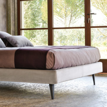 A contemporary bedroom showcases a neatly made Contemporary Upholstered Velvet Bed with a purple and brown duvet and matching pillows. Large windows behind the bed allow natural light to illuminate the room, revealing an outside view of green trees. The floor is adorned with a textured, neutral-toned rug.