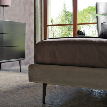 Modern bedroom interior with a large Contemporary Upholstered Velvet Bed, dark brown and beige bedding, and a black nightstand with a lamp. The space features a concrete floor, a large window with greenery visible outside, and soft natural light enhancing the room's minimalist design.