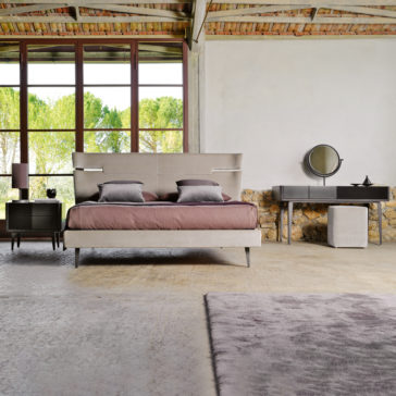 In this modern bedroom, a rustic wooden ceiling and large windows provide a view of the lush green landscape. The space is anchored by a Contemporary Upholstered Velvet Bed adorned with purple bedding. Flanking the bed are two bedside tables with lamps, while a vanity table with a mirror and an ottoman offer additional functionality. The room is tied together by its concrete floor and gray rug.