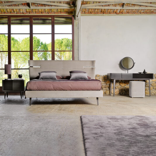 In this modern bedroom, a rustic wooden ceiling and large windows provide a view of the lush green landscape. The space is anchored by a Contemporary Upholstered Velvet Bed adorned with purple bedding. Flanking the bed are two bedside tables with lamps, while a vanity table with a mirror and an ottoman offer additional functionality. The room is tied together by its concrete floor and gray rug.