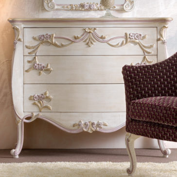 Elegant vintage wooden dresser with intricate floral carvings and gold accents. The dresser features three spacious drawers and sits on curved legs. A burgundy woven chair with white legs partially appears on the right side, all set on a light beige carpet.