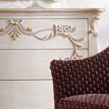 A close-up of a vintage dresser with intricate floral carvings on an ornate drawer, painted in a distressed, off-white finish with hints of pink and gold. In front of it is part of a cushioned chair upholstered in a dark red patterned fabric.