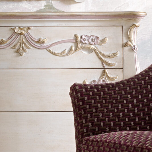 Carved Italian Chest Of Drawers A close-up of a vintage dresser with intricate floral carvings on an ornate drawer, painted in a distressed, off-white finish with hints of pink and gold. In front of it is part of a cushioned chair upholstered in a dark red patterned fabric.