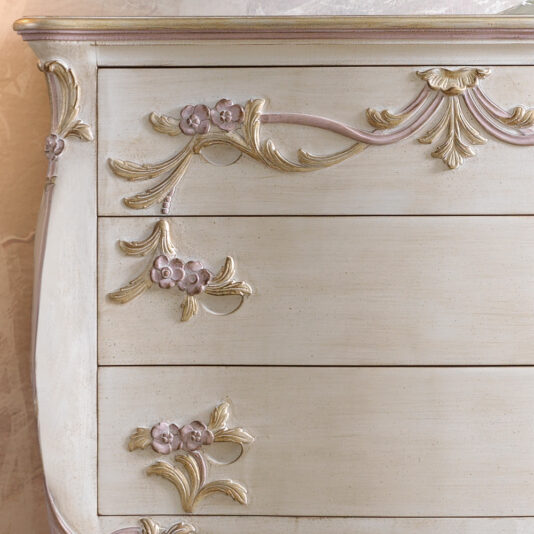 Carved Italian Chest Of Drawers Close-up of an ornate antique dresser with three visible drawers. The dresser is painted in a soft cream color and features floral carvings with pastel pink and gold accents, adding a delicate and vintage touch to the furniture.
