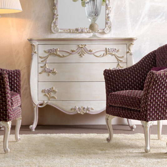 An elegant room features two upholstered chairs with maroon and white woven fabric, positioned on either side of a vintage white chest of drawers with ornate floral carvings. A large mirror with a decorative frame hangs above the chest, and a lamp is visible to the left.