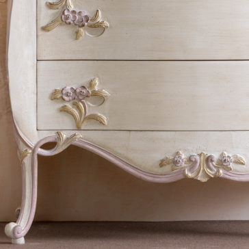 An ornate vintage dresser with a carved, curved design. The drawers feature floral decorations in soft pink and gold accents, adding an elegant touch to the antiqued white finish. The dresser stands on slender, gracefully curved legs with intricate detailing.