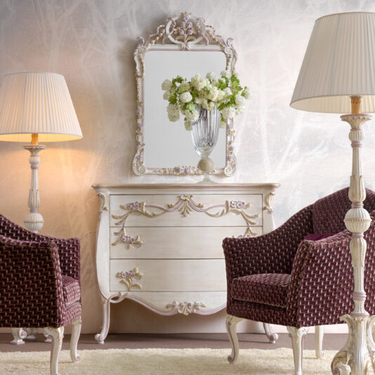 Carved Italian Chest Of Drawers Elegant room featuring a decorative white dresser with ornate floral details and a matching mirror, flanked by two cream-colored lamps with pleated shades. In front are two upholstered chairs with a dark woven pattern and intricate, carved legs. A vase of white flowers sits atop the dresser.