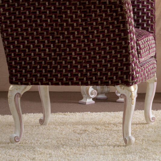 An ornate Italian Carved Upholstered Armchair, featuring a woven maroon and gold checkered fabric design on the seat and backrest, along with elegant white wooden legs adorned with intricate carvings, is positioned on a plush, light-colored carpet. Another armchair of similar design is partially visible in the background.
