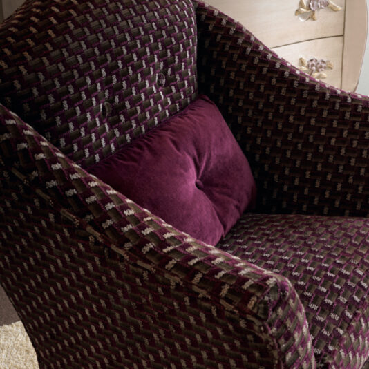 The Italian Carved Upholstered Armchair is a plush seat adorned in dark purple fabric with a checkerboard design, complemented by a deep purple velvety pillow. It stands against a light-colored background with a glimpse of light wood dresser visible in the corner.