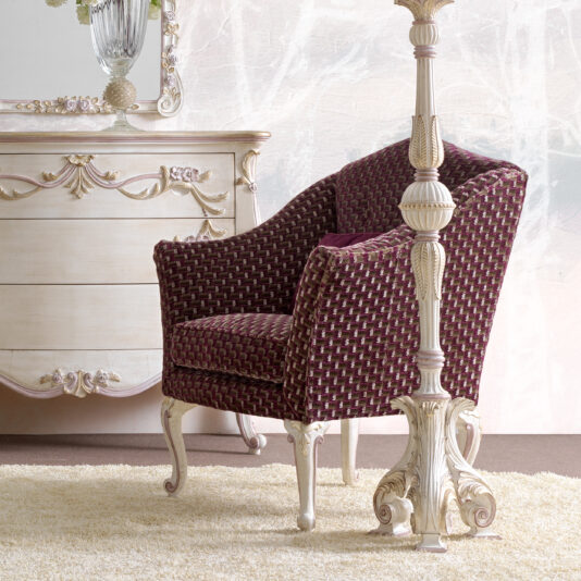 A plush Italian Carved Upholstered Armchair in burgundy, showcasing intricate patterned upholstery, is positioned next to an ornate cream-colored dresser adorned with detailed carvings. These elegant furnishings rest on a light beige carpet, presenting a sophisticated and classic interior design.