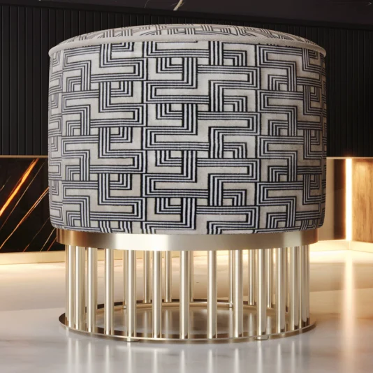 The Luxury Pouffe With Brass Base showcases a black and white geometric pattern on top and vertical brass bars on the base, bringing modern elegance. When placed on a white marble floor, it transforms into an opulent centerpiece for any room.