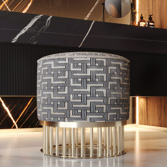 The Luxury Pouffe with Brass Base features vertical bars and stands on a marble floor in a stylish, well-lit room accented with dark wood. This cylindrical stool boasts a maze-like geometric black-and-white design on its fabric upholstery, adding modern luxury to the space.