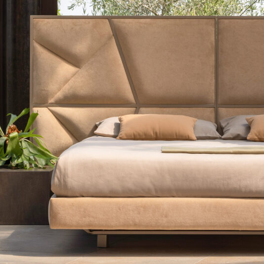 A modern bedroom showcasing the Modern Abstract Upholstered Bed, featuring a plush, beige design with a rectangular and geometrically designed headboard. The bed is adorned with matching beige pillows and a neatly folded blanket. A small green potted plant sits on the side, enhancing the room's ambiance. Natural light floods the space, highlighting its contemporary charm.