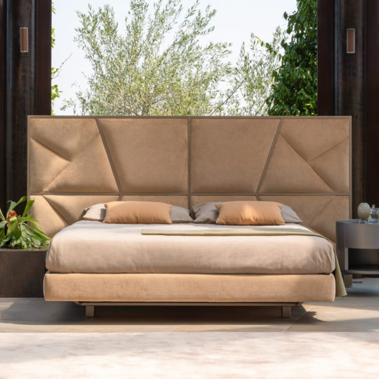 A modern, neutral-toned bedroom features the Modern Abstract Upholstered Bed with a large, geometrically designed beige headboard. The bed is complemented by neatly arranged pillows and bedding. Tall windows behind the bed reveal lush green foliage and clear skies, while a round side table nearby enhances the modern abstract aesthetic.