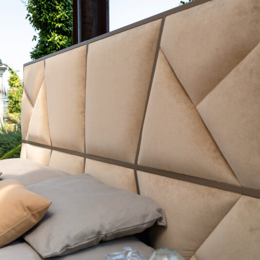 A close-up view of the Modern Abstract Upholstered Bed showcases its geometric beige headboard adorned with triangular and rectangular shapes. The bed is impeccably arranged with beige and light grey bedding, while greenery and a portion of an outdoor area are visible in the background, further enhancing the serene ambiance.