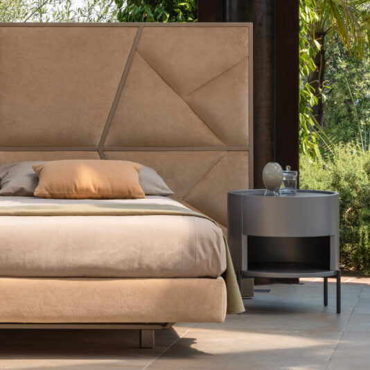 A contemporary bedroom showcases the Modern Abstract Upholstered Bed with a chic beige quilted headboard, complemented by matching bedding. A mustard yellow pillow provides a vibrant touch of color. Next to the bed, a round gray nightstand supports a lamp and a small dish. Expansive windows reveal lush greenery outside, offering an abstract view of nature.